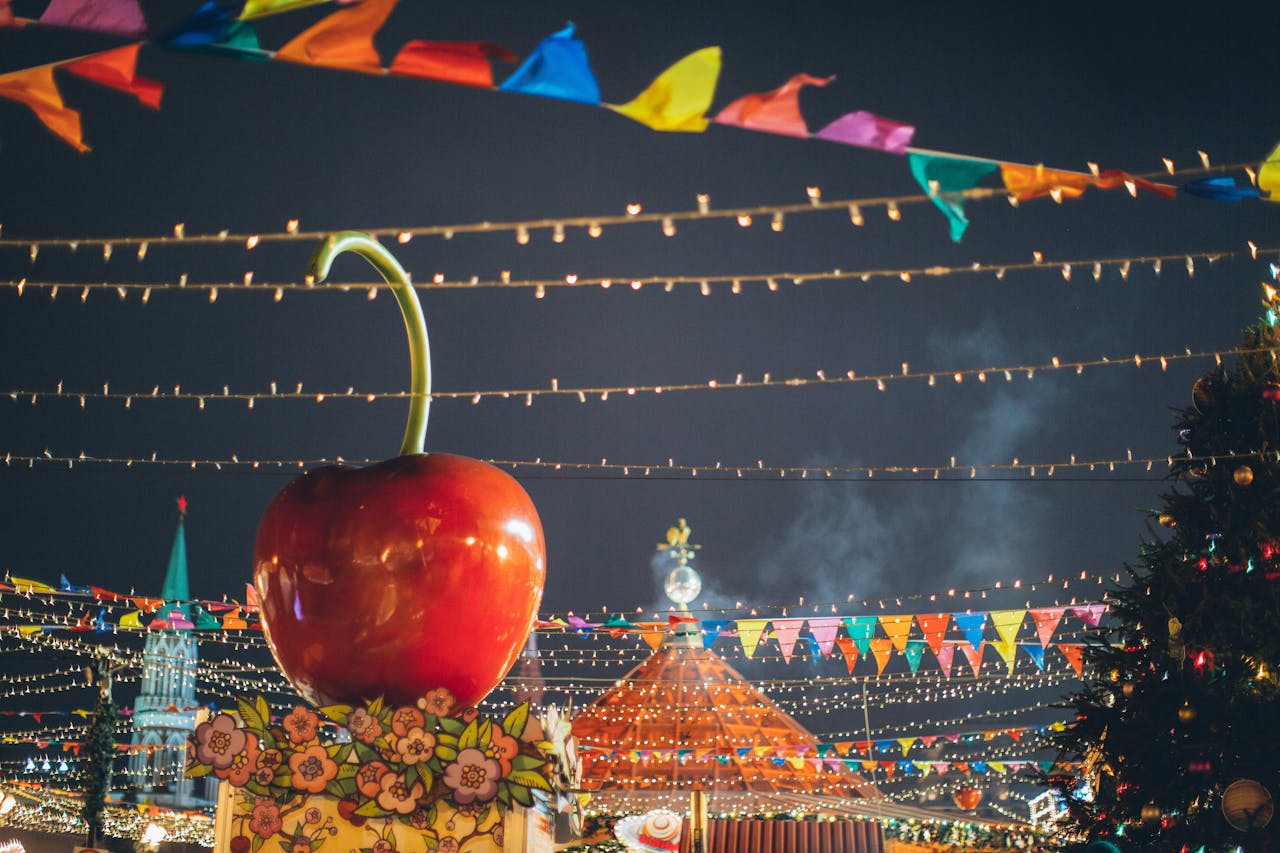 Big red glossy toy apple on roof of building on fairground against dark sky in evening city park decorated to winter holidays