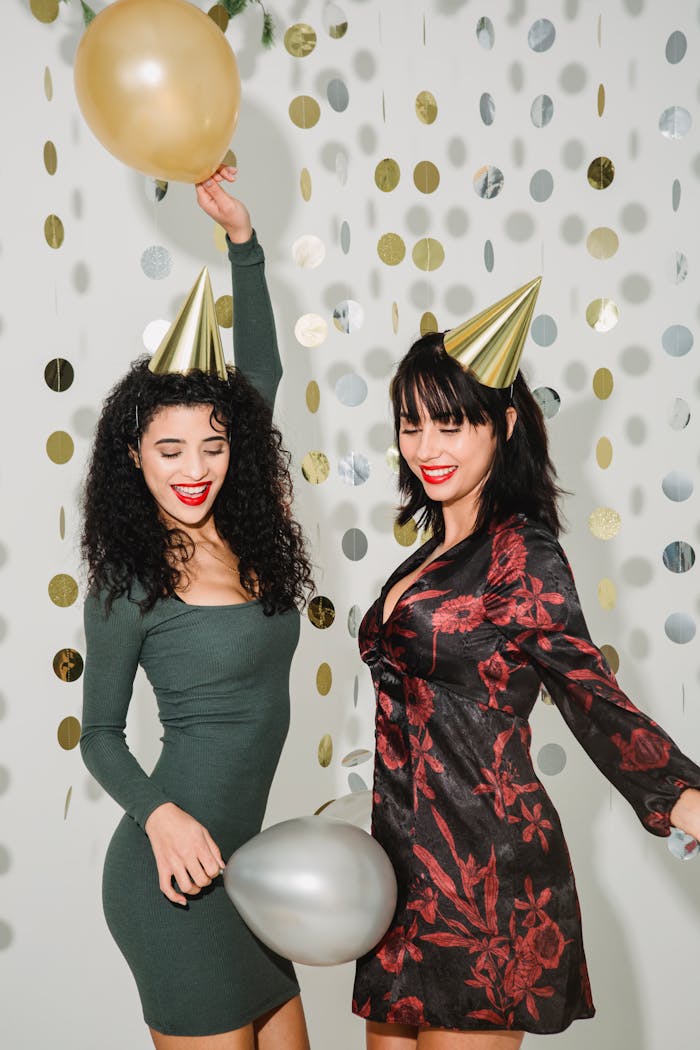 Happy female friends with helium balloons wearing party hats having fun while standing on white background with paper garland during holiday celebration